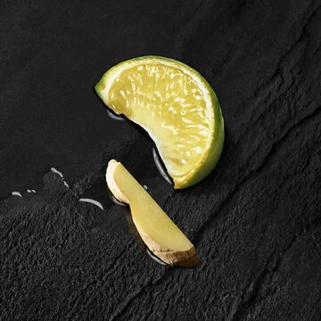 Tranche de citron vert et morceau de gingembre sur une surface noire, avec quelques gouttes de jus.