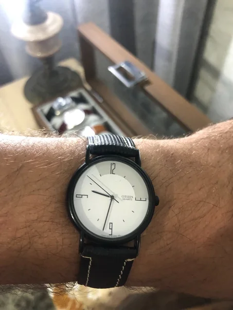 minimalist Citizen Quartz watch featuring a clean white dial, black matte case, and a textured black fabric or canvas strap.